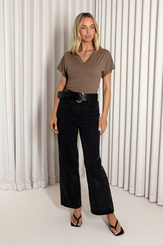 A woman stands in front of white pleated curtains wearing a taupe brown v-neck t-shirt, black wide-leg corduroy pants with a black belt, and black strappy heels.
