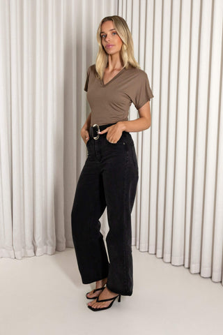 A woman modeling a taupe brown short-sleeve V-neck top and black wide-leg jeans with a silver belt, standing against a white pleated curtain backdrop.