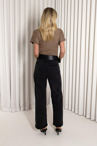 A woman viewed from behind wearing a brown short-sleeve top, a black leather belt, black wide-leg jeans, and black high heels, standing in front of a white pleated curtain backdrop.