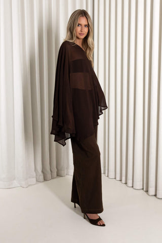 A woman modeling a dark brown sheer and opaque two-piece outfit consisting of a draped long-sleeve top and matching wide-leg trousers, standing against a white pleated curtain backdrop.