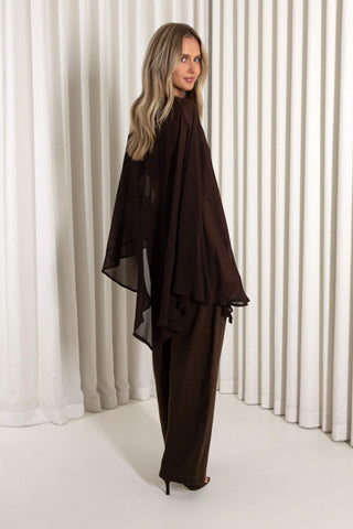 A woman modeling a dark brown two-piece outfit consisting of a sheer draped long-sleeve top and matching wide-leg trousers, standing in front of white vertical drapes.