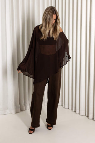A woman modeling a dark brown two-piece outfit consisting of a sheer off-the-shoulder long-sleeve top and matching wide-leg trousers, standing in front of white vertical drapes.