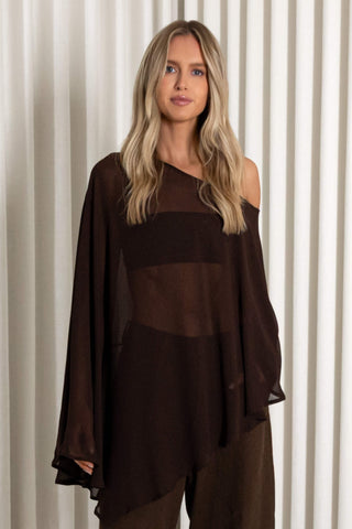 A woman with blonde hair wearing a dark brown sheer and opaque asymmetrical long-sleeve top, paired with matching brown pants, standing in front of a white pleated curtain background.
