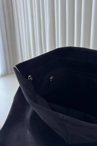 Close-up of the interior opening of a black leather or faux-leather bag, showing the dark interior lining and small silver zipper pulls, with a softly blurred white curtain background.
