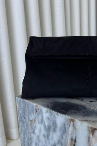 A black suede or nubuck clutch bag with a folded-over top flap, resting on a gray marble block in front of white curtains.