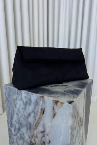 A black fabric clutch bag with a fold-over flap, displayed on a white and gray marble pedestal in front of white pleated curtains.