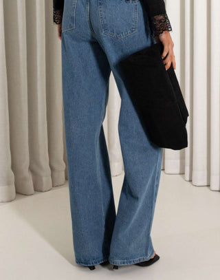 Person wearing blue jeans holding a black clutch against a neutral background