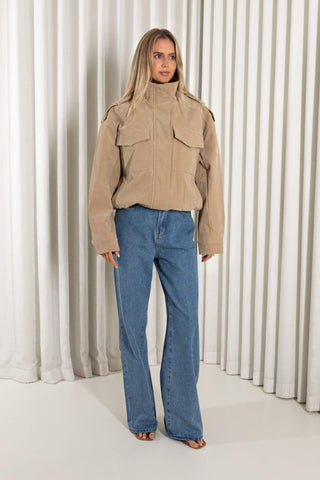 A woman modeling a beige jacket with flap chest pockets and blue wide-leg jeans, standing in front of a white pleated curtain backdrop.
