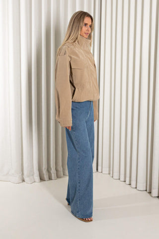 A woman stands in profile wearing a beige jacket and wide-leg blue jeans against a studio backdrop with vertical white drapes.