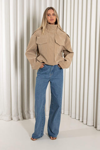 A woman modeling a beige jacket with shoulder epaulets and front flap pockets, paired with wide-leg blue jeans, standing in front of a white pleated curtain backdrop.
