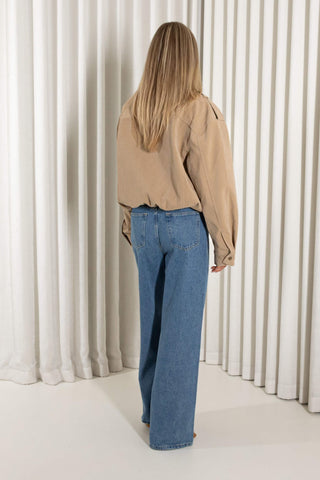 A woman with long blonde hair is shown from behind wearing a beige long-sleeve button-up jacket and wide-leg blue jeans, standing in front of a white pleated curtain backdrop