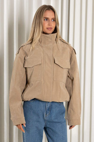 A woman wearing a cropped beige faux suede jacket with a high stand-up collar and flap chest pockets, paired with blue jeans, standing against a white pleated curtain background.
