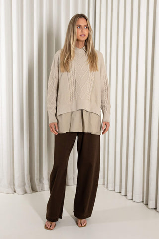 A woman stands in heels in front of white vertical drapes, wearing a cream cable-knit mock-neck sweater over a matching beige peplum-style top and dark brown wide-leg pants.