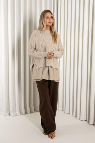 A woman stands in heels in front of white vertical drapes, wearing a cream cable-knit mock-neck sweater over a matching beige peplum-style top and dark brown wide-leg pants.