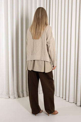 A woman stands in heels in front of white vertical drapes, wearing a cream cable-knit mock-neck sweater over a matching beige peplum-style top and dark brown wide-leg pants.