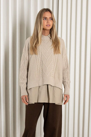 A woman stands in heels in front of white vertical drapes, wearing a cream cable-knit mock-neck sweater over a matching beige peplum-style top and dark brown wide-leg pants.