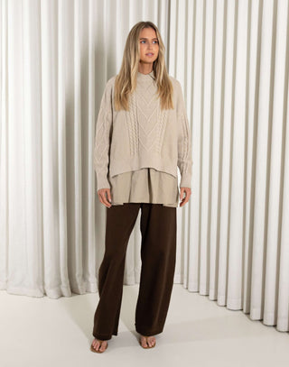 Woman wearing a beige cable knit sweater and brown pants against a white curtain background