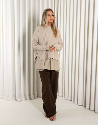 Woman wearing a beige cable knit sweater and brown pants standing against a white curtain.