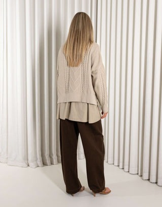 Person wearing a beige cardigan and brown pants standing in front of white curtains.