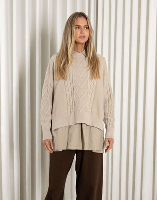 Woman wearing a beige cable knit sweater and brown pants against a neutral background