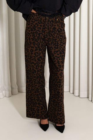 A person wearing brown and black leopard print wide-leg pants, paired with a black top and black pointed-toe heels, standing in front of white curtains.