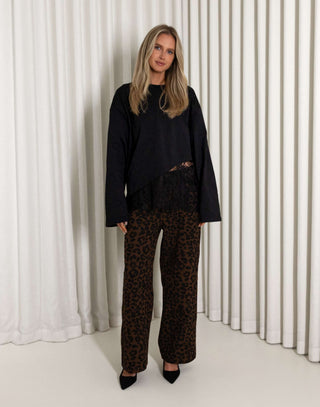 Woman wearing a black lace top and leopard print pants standing against a white curtain.