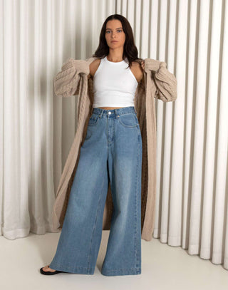 Woman wearing a long nude cardigan, white top, and blue jeans against a white curtain background nude longline cable knit cardigan