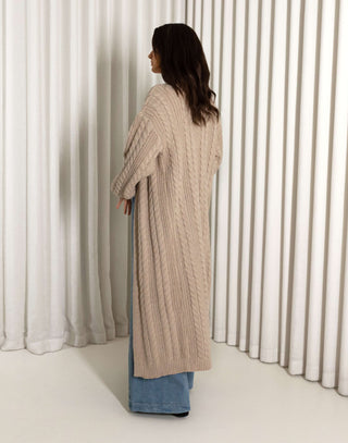 Woman wearing a long nude cardigan, white top, and blue jeans against a white curtain background nude longline cable knit cardigan