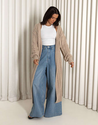 Woman wearing a long nude cardigan, white top, and blue jeans against a white curtain background nude longline cable knit cardigan