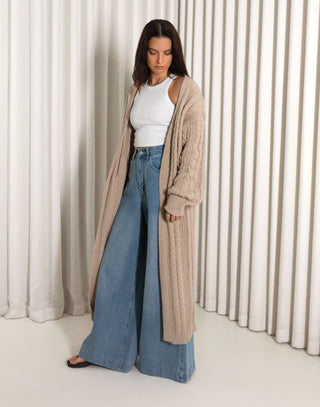 Woman wearing a long nude cardigan, white top, and blue jeans against a white curtain background nude longline cable knit cardigan