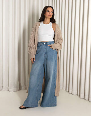 Woman wearing a long nude cardigan, white top, and blue jeans against a white curtain background nude longline cable knit cardigan
