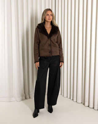 Woman wearing a brown jacket with fur collar and black pants against a white curtain background
