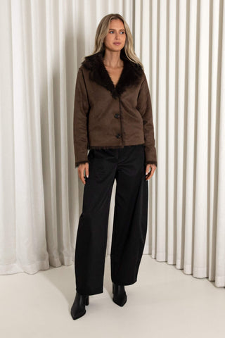 A woman modeling a dark brown reversible faux fur jacket with black wide-leg trousers and black pointed-toe boots, standing in front of a white curtain backdrop.