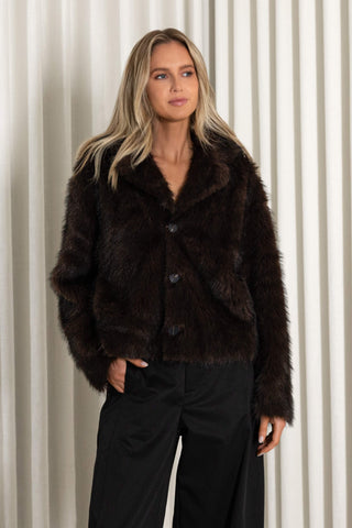 A woman modeling a dark brown reversible faux fur jacket with black wide-leg trousers and black pointed-toe boots, standing in front of a white curtain backdrop.
