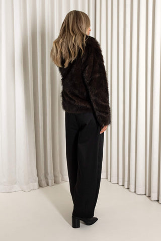 A woman modeling a dark brown reversible faux fur jacket with black wide-leg trousers and black pointed-toe boots, standing in front of a white curtain backdrop.