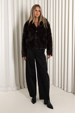 A woman modeling a dark brown reversible faux fur jacket with black wide-leg trousers and black pointed-toe boots, standing in front of a white curtain backdrop.