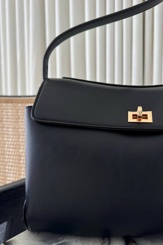 A close-up of a black leather handbag with a gold turn-lock clasp and a curved shoulder strap, set against a background of white vertical blinds and a woven chair.