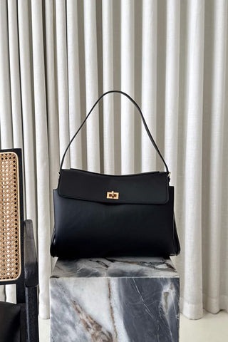 A black leather shoulder bag with a gold-tone turn-lock clasp, displayed on a white and gray marble pedestal in front of white pleated curtains.