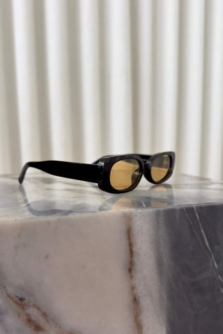 Black square sunglasses with yellow-tinted lenses resting on a polished white and gray marble block against a soft beige curtain background.