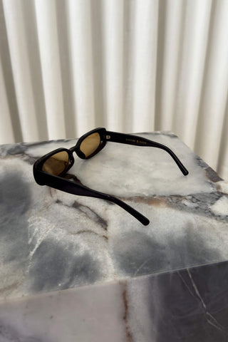 Black square sunglasses with yellow-tinted lenses resting on a gray and white marble block in front of a white pleated curtain background.