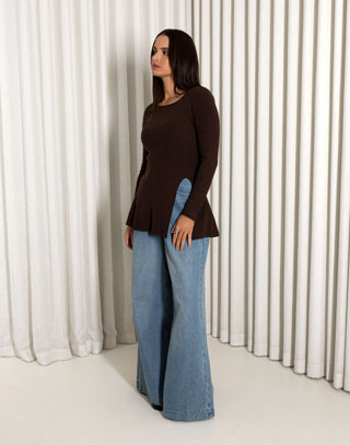 Woman wearing a brown top and blue jeans standing against a white curtain background
