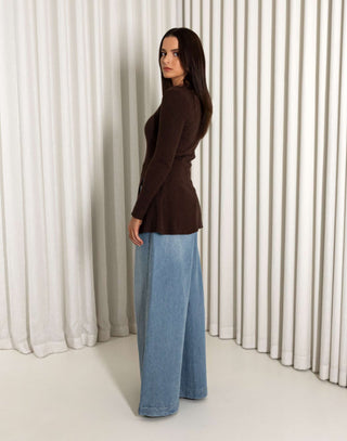 Woman wearing a brown top and blue jeans standing against a white curtain background