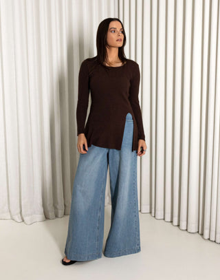Woman wearing a brown top and blue jeans standing against a white curtain background