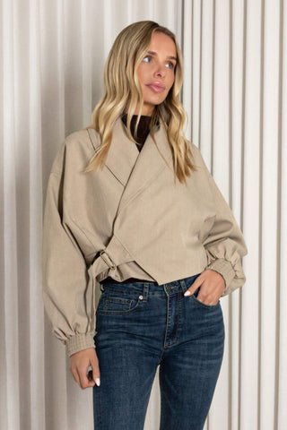 A woman wearing a beige wrap-style jacket with balloon sleeves and dark blue jeans, standing in front of a white pleated curtain backdrop.