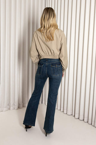 A woman viewed from behind wearing a beige cropped bomber-style jacket and dark blue flare jeans, standing in front of a white pleated curtain backdrop.