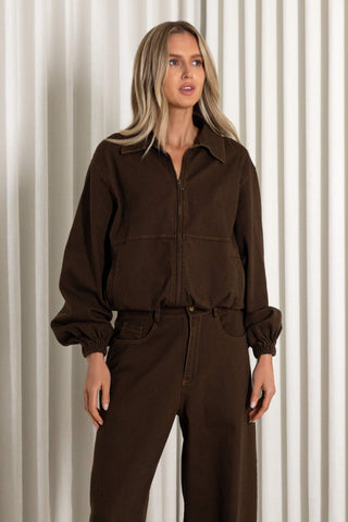 A woman modeling a dark brown zip-up utility jacket with matching wide-leg trousers, standing against a background of vertical beige drapes.