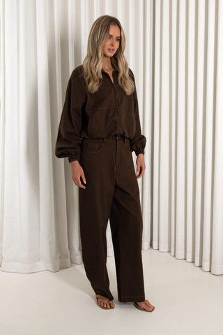 A woman modeling a dark brown long-sleeve button-up shirt and matching wide-leg trousers set, standing against a white curtain backdrop.