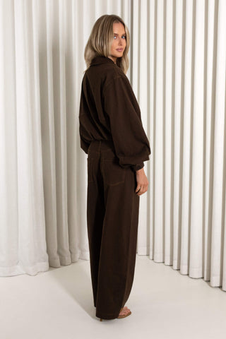 A woman stands in profile wearing a dark brown long-sleeve jumpsuit with a relaxed, oversized fit, set against a background of white vertical drapes.