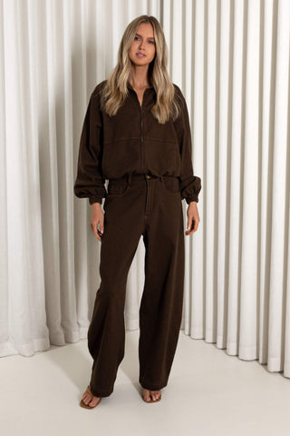 A woman modeling a dark brown corduroy jumpsuit with a button-up front, long sleeves, and wide-leg pants, standing against a white pleated curtain backdrop.