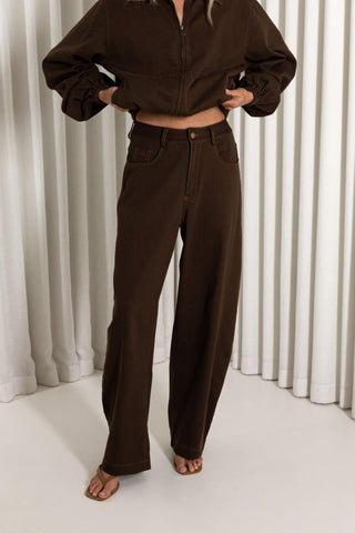 A model wearing a dark brown cropped zip-up jacket and matching wide-leg jeans, standing barefoot in front of white vertical drapes.